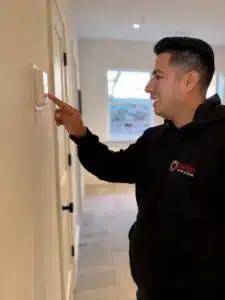 A man in a black hoodie adjusts a thermostat on a wall in a well-lit room with carpeted floors and a window in the background.